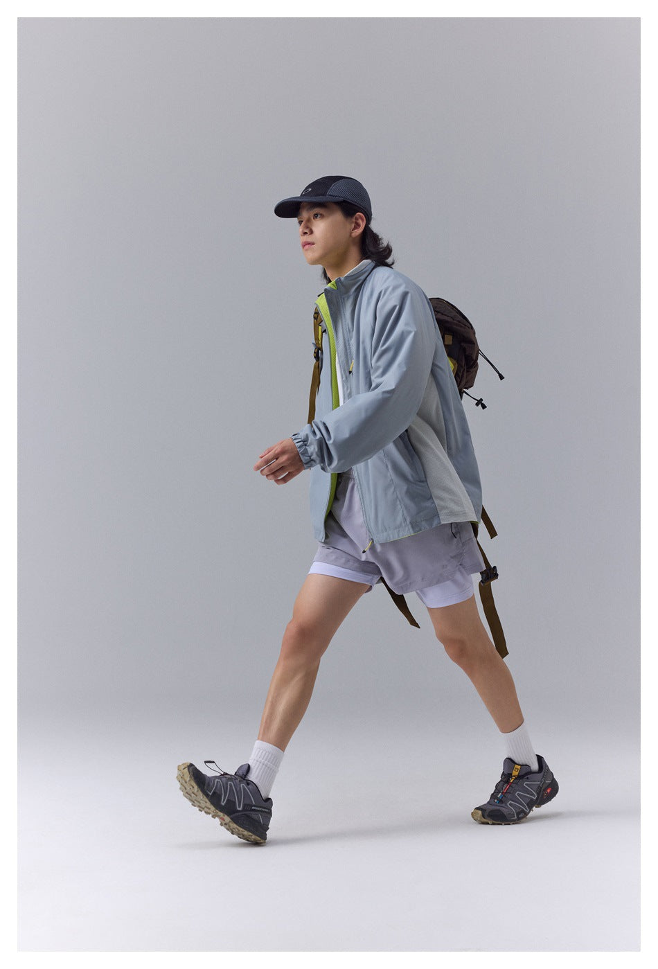 Model walking in a pale blue-gray zip jacket with lime accents, gray shorts, and a backpack.