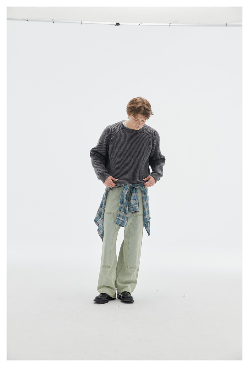 Full-length model wearing a charcoal ribbed crewneck sweater, pale green jeans, and a blue plaid shirt tied at the waist.