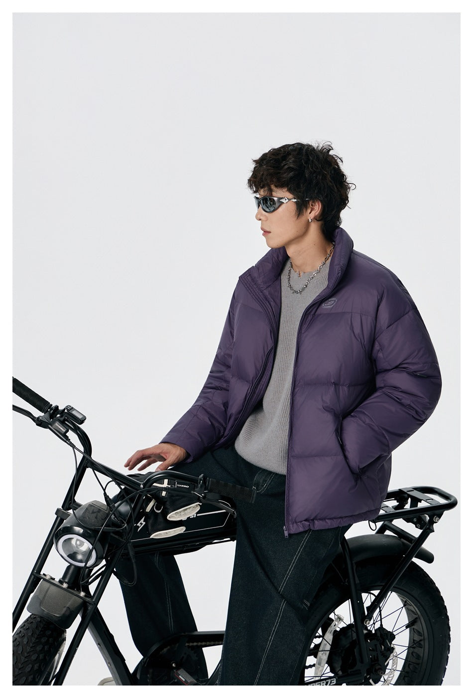 Model wearing an open purple puffer jacket while seated on a bicycle; the jacket has a high collar and side pockets.