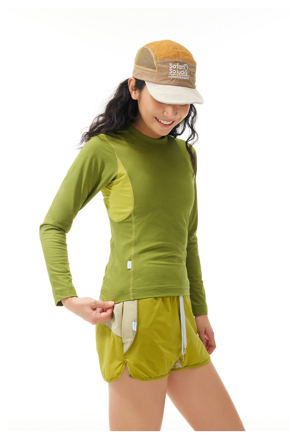 Model wearing a green long-sleeve top with mesh side panels and yellow shorts, plus a tan-and-cream cap.