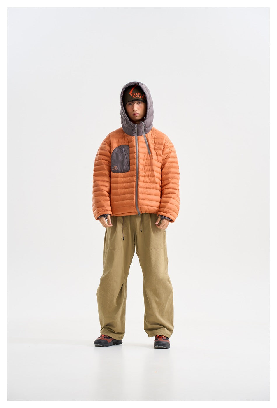 Front view of a model wearing an orange quilted hooded jacket with a gray chest pocket and loose olive trousers.