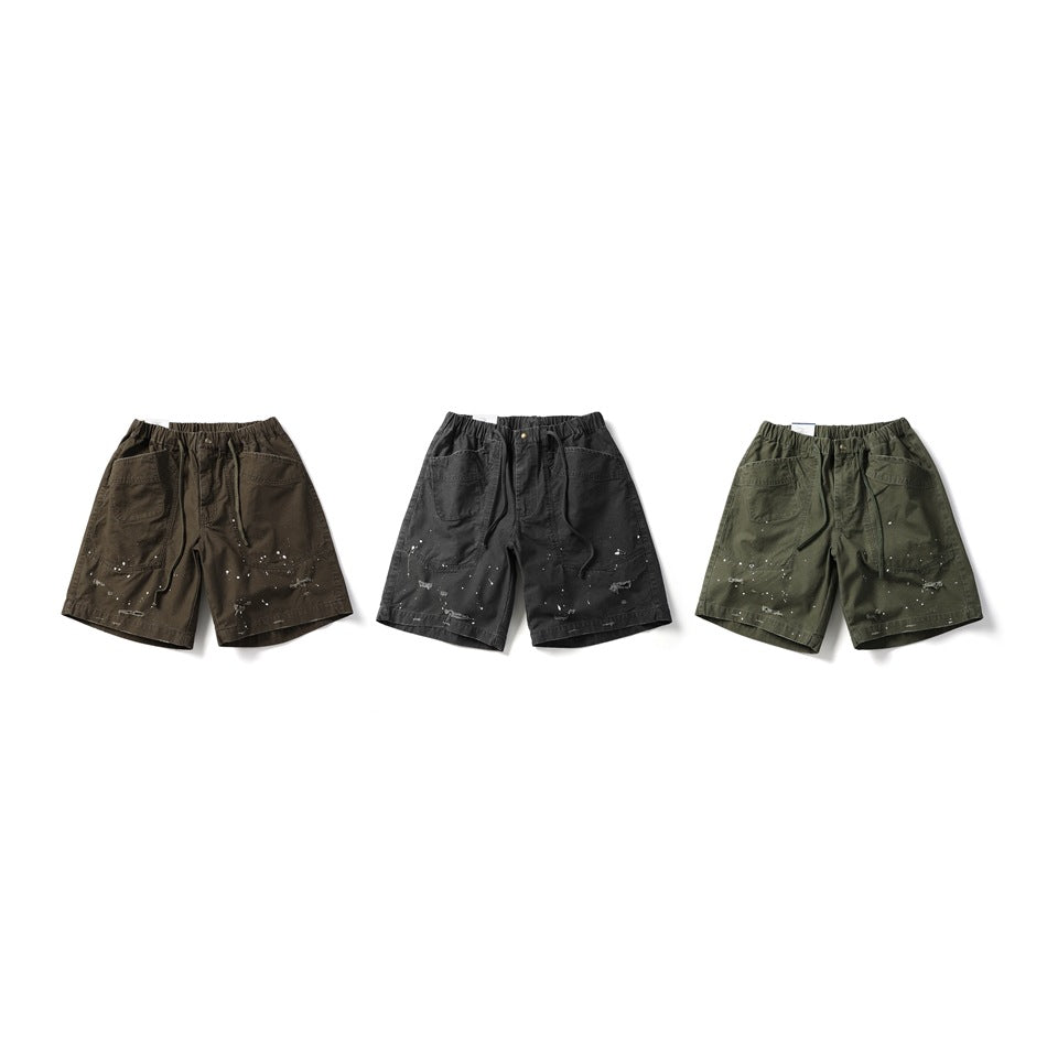 Three pairs of drawstring shorts in brown, dark gray and army green with paint splatters, laid out side by side