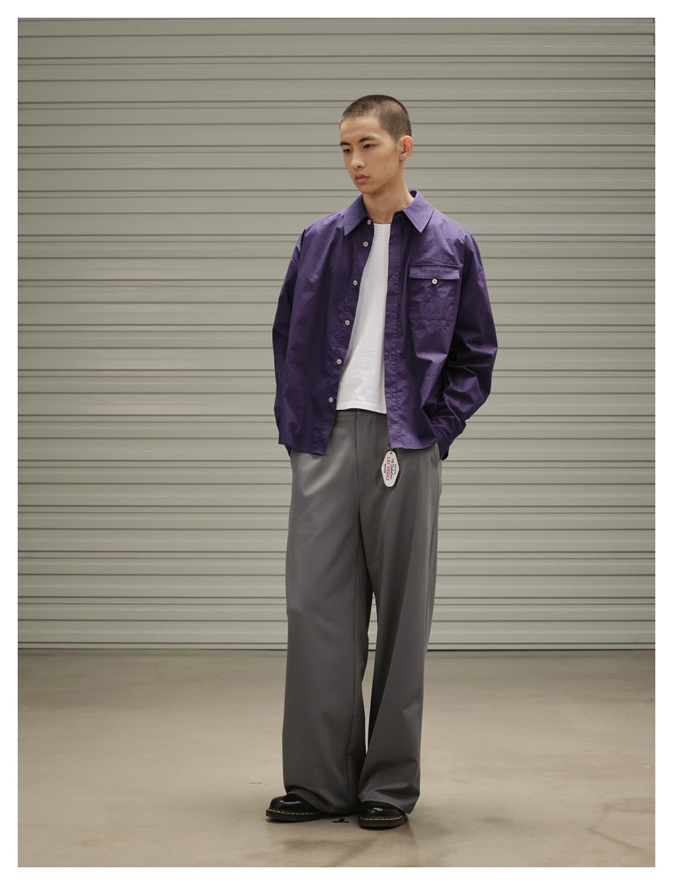 Model in a purple button-up overshirt over a white T-shirt and wide gray trousers, standing at a slight angle.