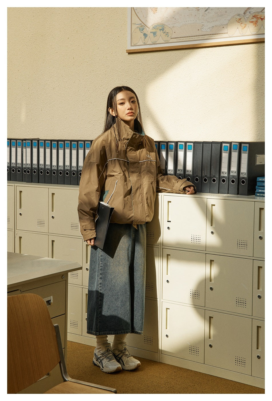 Person wearing a brown stand-collar jacket with white piping over a long denim skirt in an office-style setting