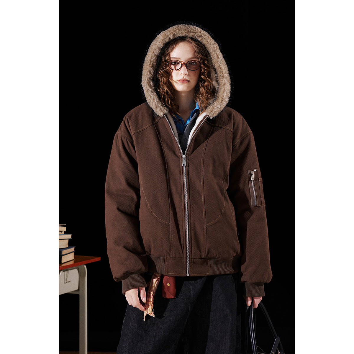 Front view of a model wearing a brown zip-up jacket with a fur-trimmed hood and a zip pocket on the sleeve.
