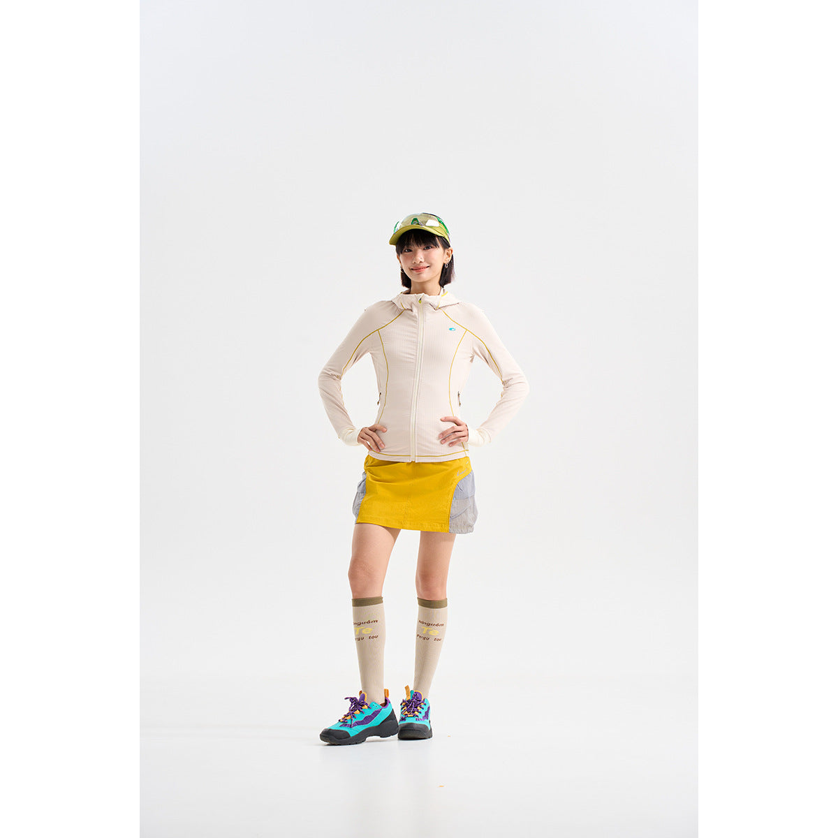Model wearing a cream zip-up jacket, yellow-and-gray skort, knee-high socks, and colorful sneakers.