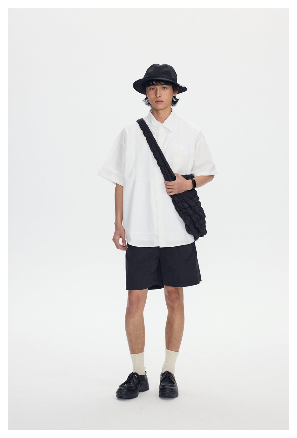 Model in a white short-sleeve button-up shirt and black shorts, carrying a textured black crossbody bag.
