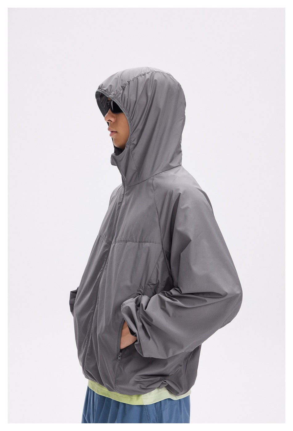 Side view of a model wearing a loose gray hooded jacket with a front zip and pockets, over blue bottoms.