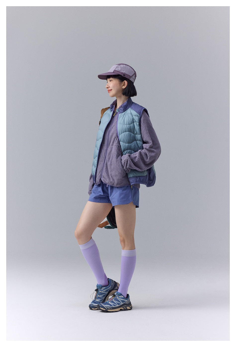 Side view of a model wearing a pale blue quilted vest with purple trim, a textured purple top and blue shorts.