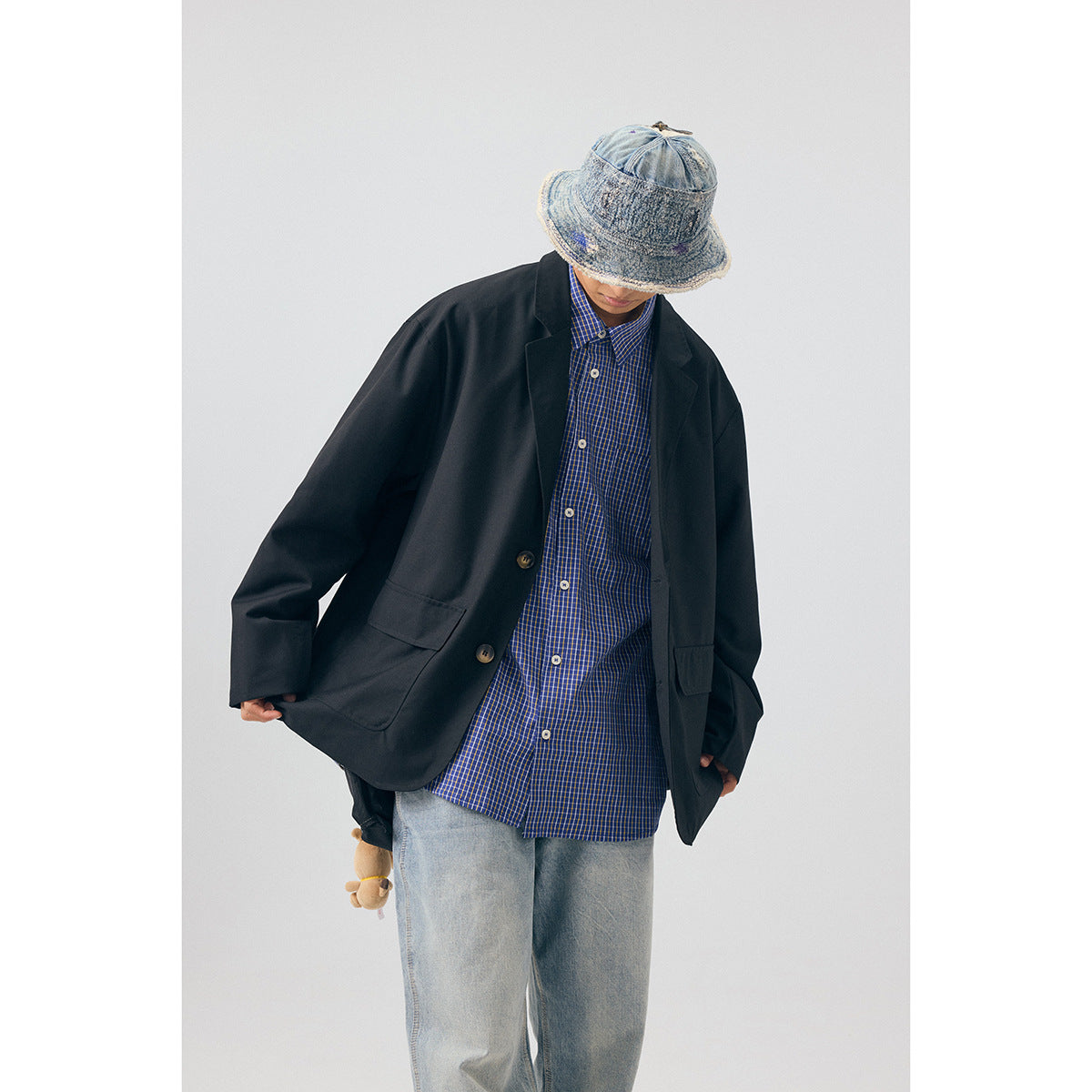 Model wearing an oversized black blazer over a blue checked shirt, light jeans and a denim bucket hat; front view.