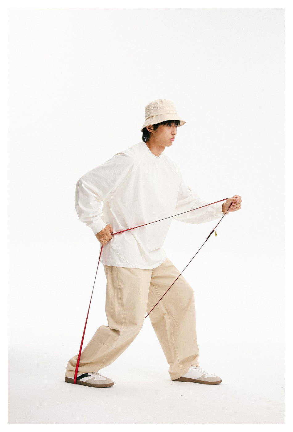 Side view of a model wearing a loose white long-sleeve shirt, beige trousers, and bucket hat while holding a red strap.