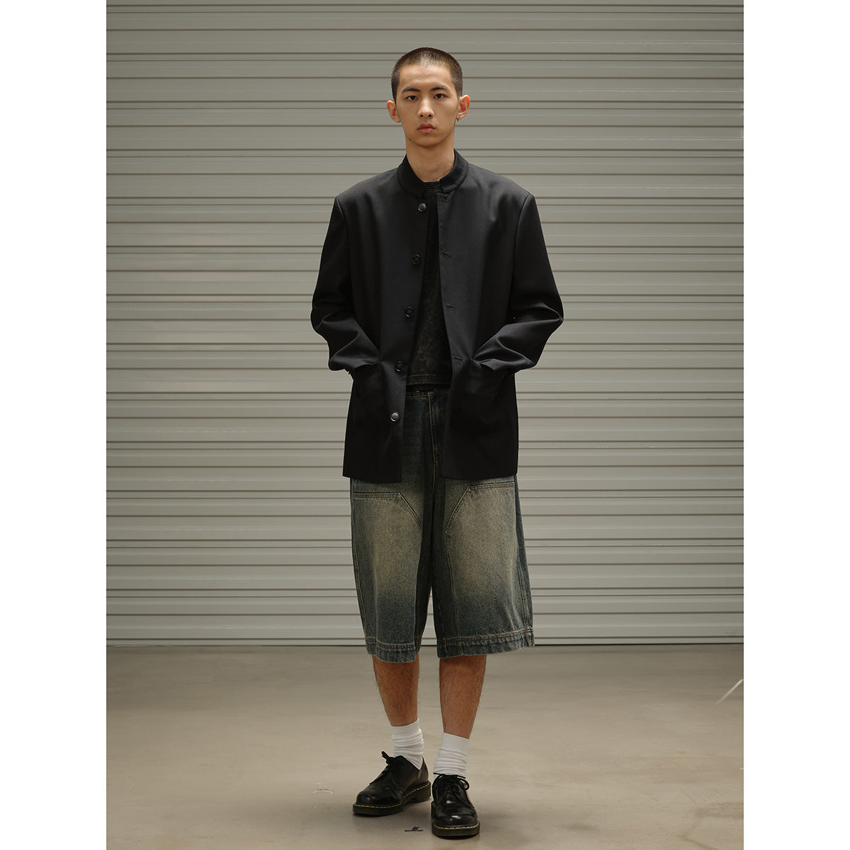 Full-length model wearing a black band-collar button coat, dark top, oversized faded denim shorts, white socks and black