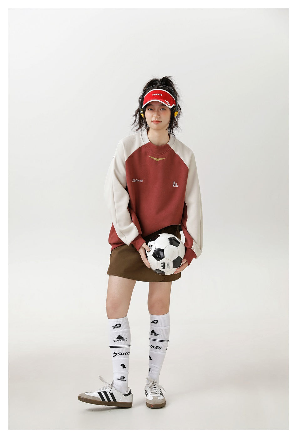 A model wears a rust-and-cream raglan sweatshirt with a brown skirt, holding a soccer ball.