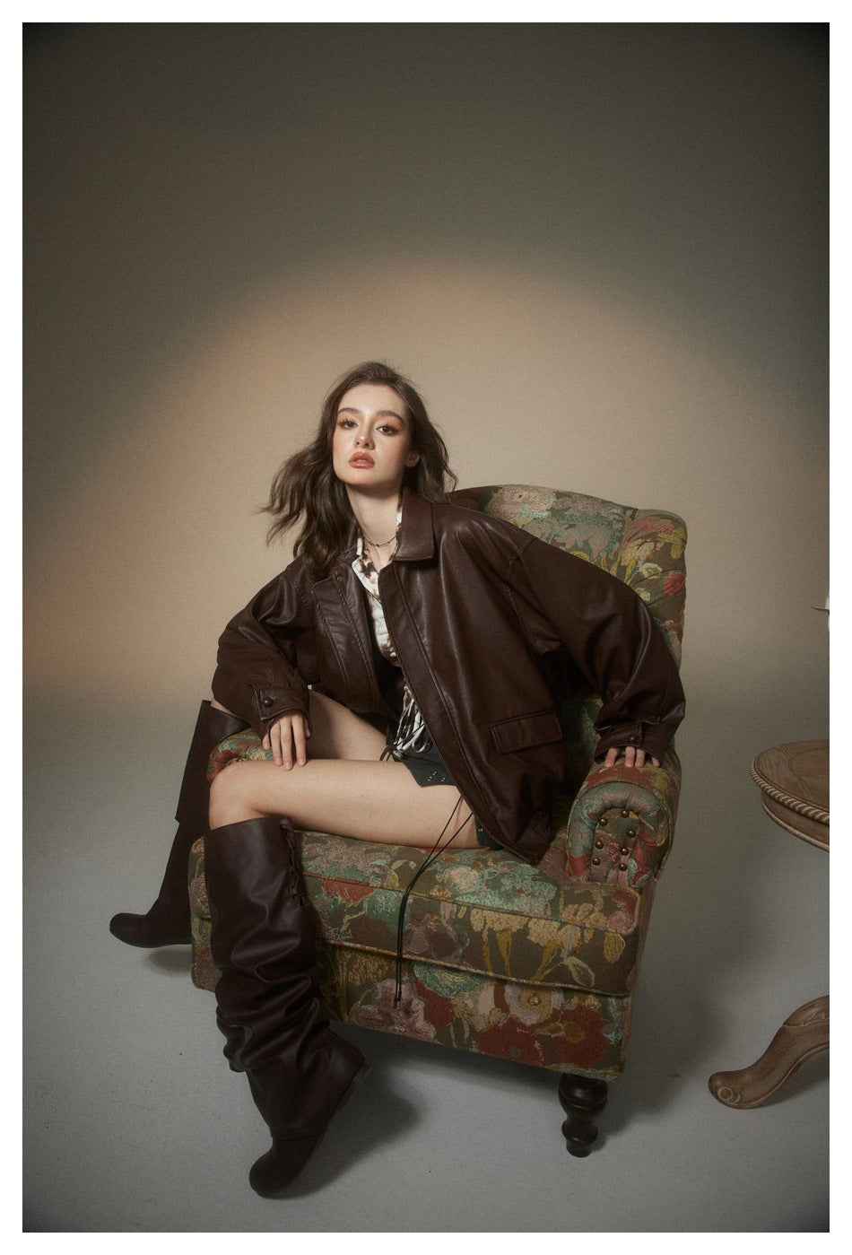 Model seated in an oversized dark brown jacket over a printed top, with a black mini skirt and tall boots.