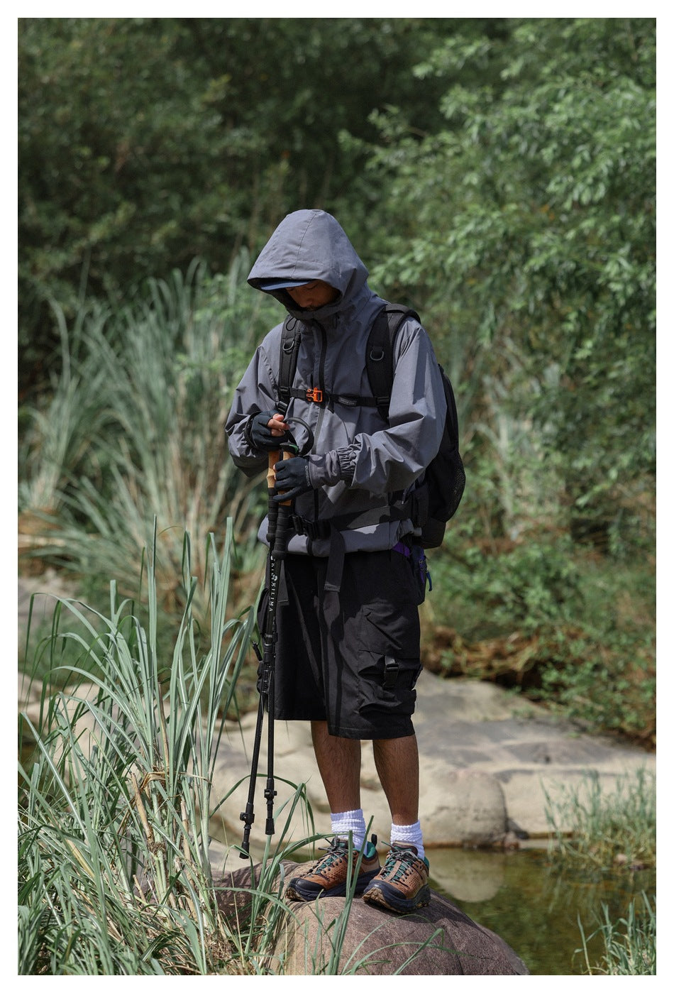 Man wearing a gray hooded jacket and black cargo shorts while holding trekking poles on a rocky trail.