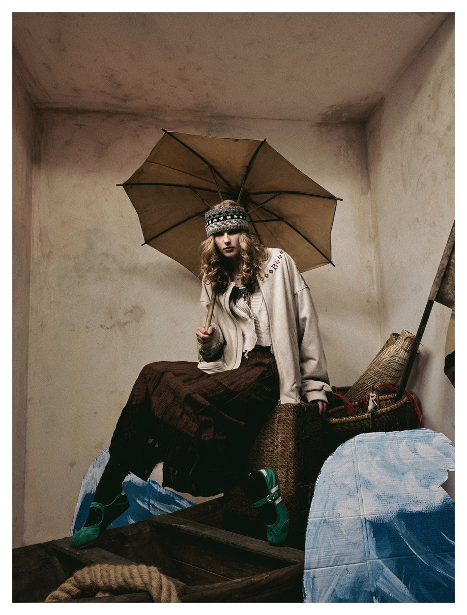 Model seated beneath an umbrella wearing an ivory embroidered jacket, dark skirt, and patterned headband.