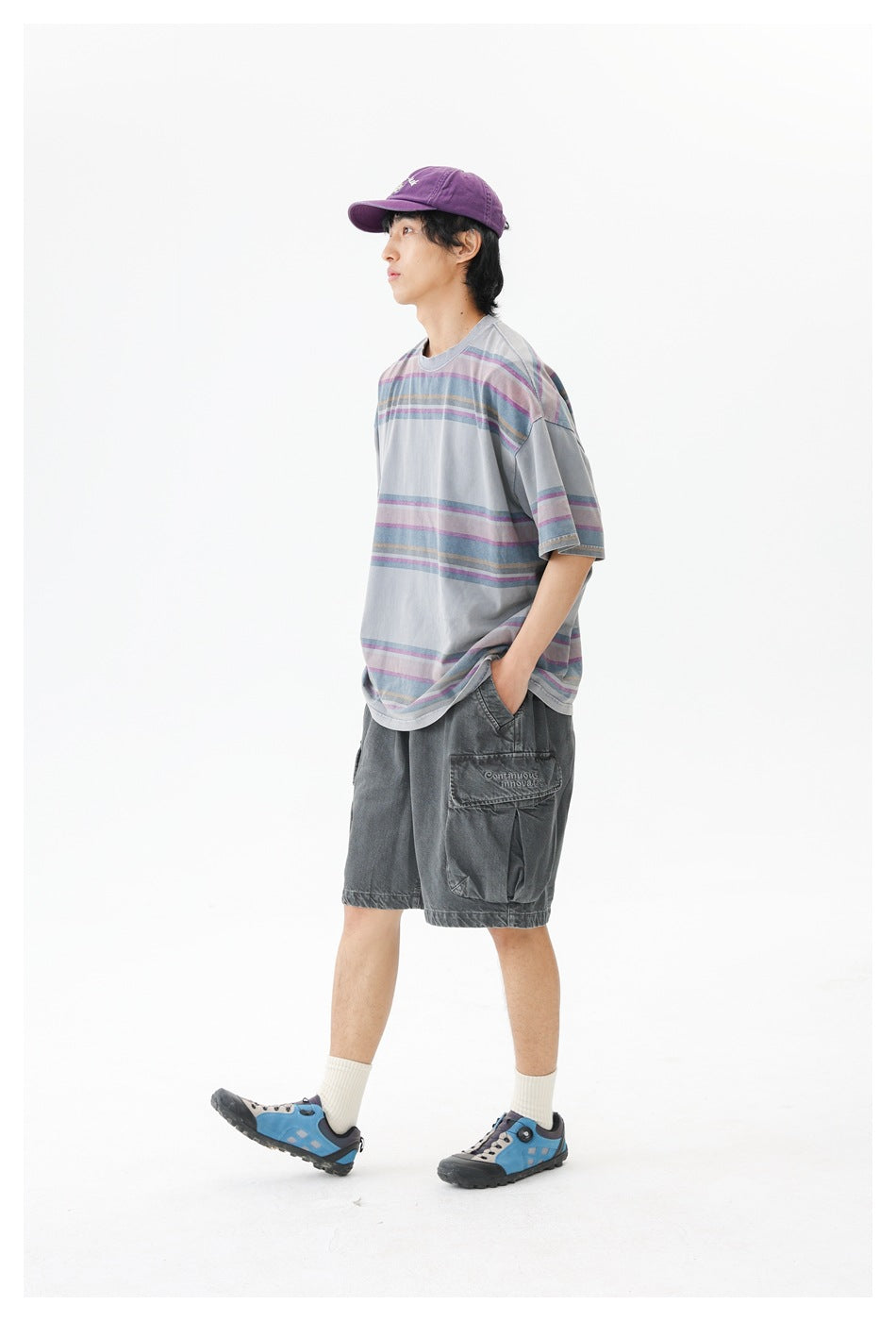 Model in a loose gray T-shirt with muted purple, blue, and tan horizontal stripes, charcoal cargo shorts, and a purple cap.