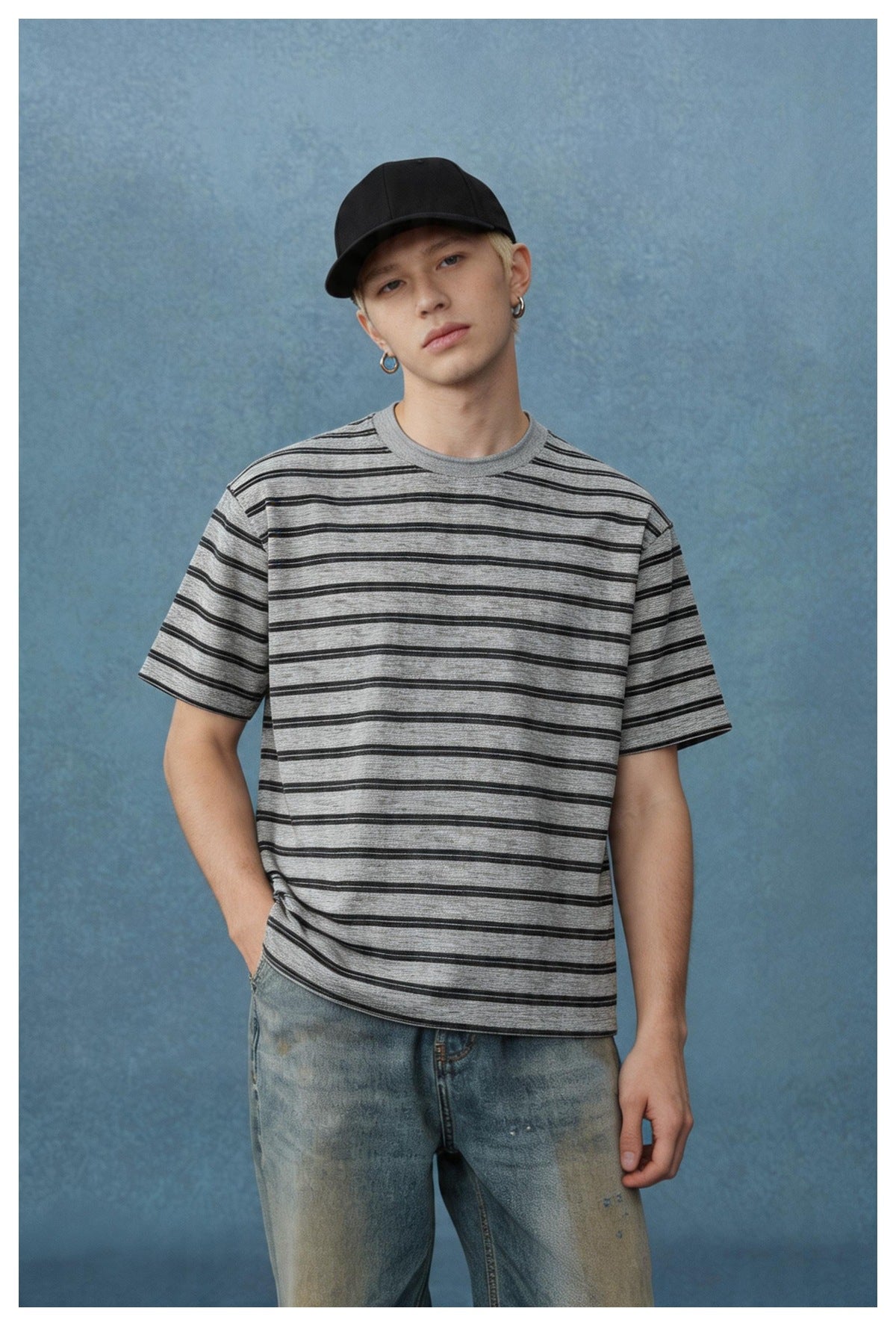 Model wearing a black and gray striped short-sleeve T-shirt with a black cap and faded jeans against a blue backdrop