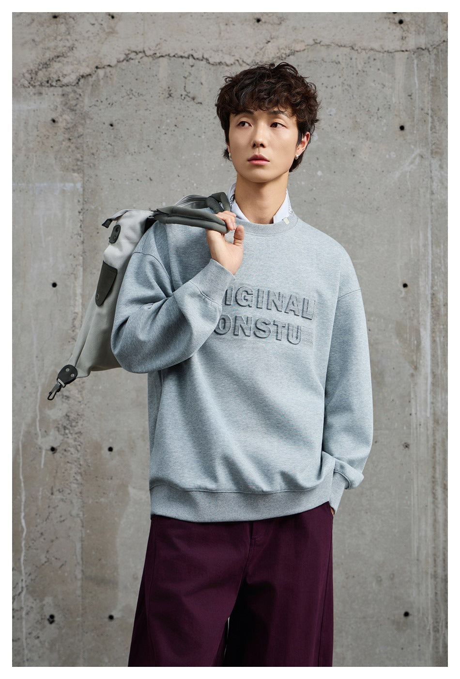 Model wearing gray crewneck sweatshirt with embossed chest lettering, holding a bag over the shoulder against concrete