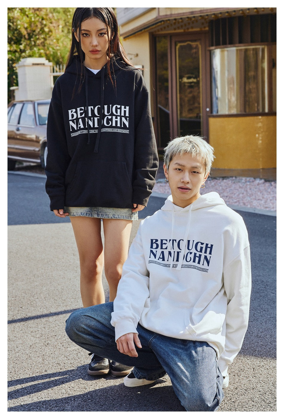 Two models wear oversized graphic hoodies, one black and one white, styled with denim bottoms outdoors.
