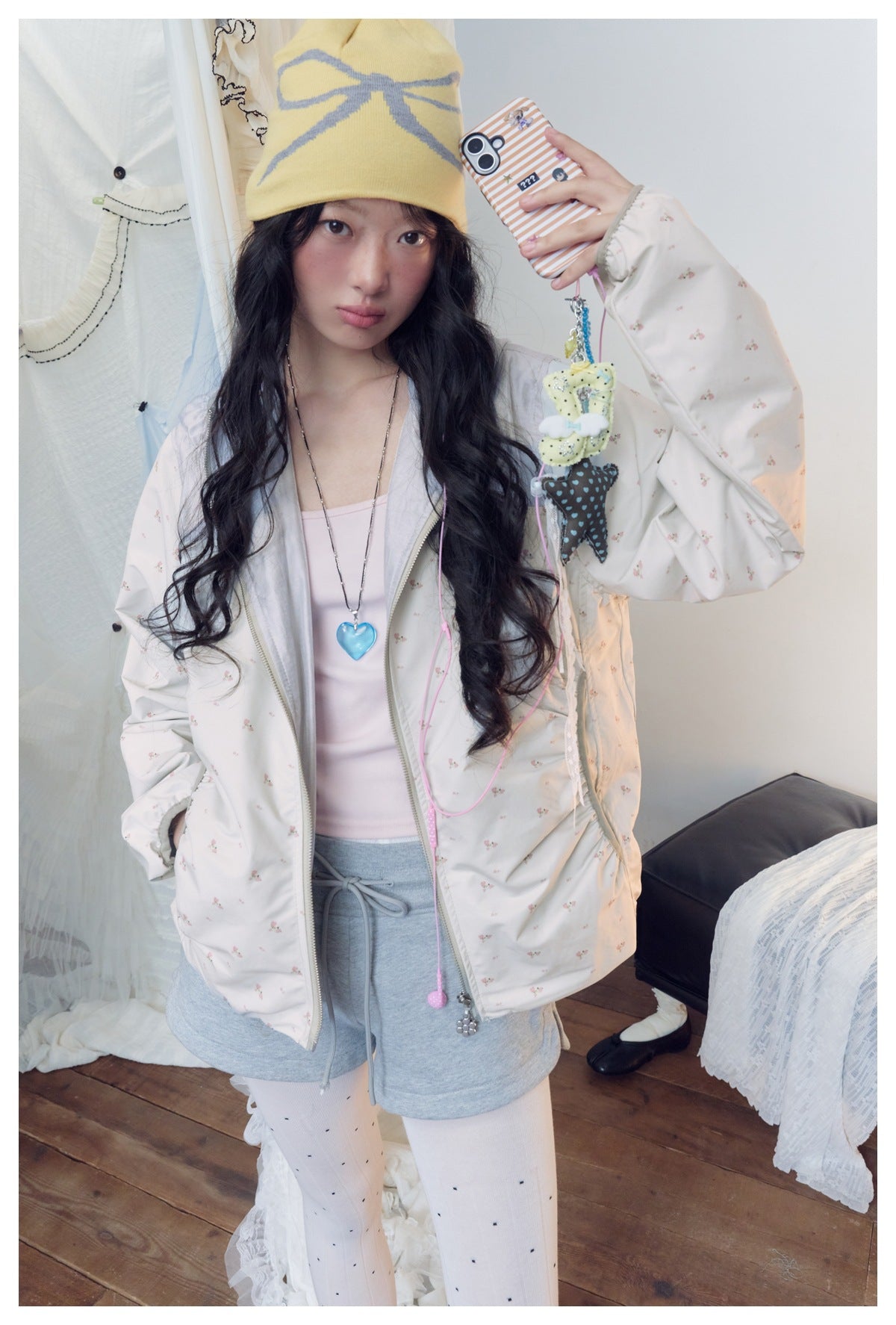 Closer front view of a model in a pale floral zip jacket with gray shorts and a yellow bow-pattern beanie.