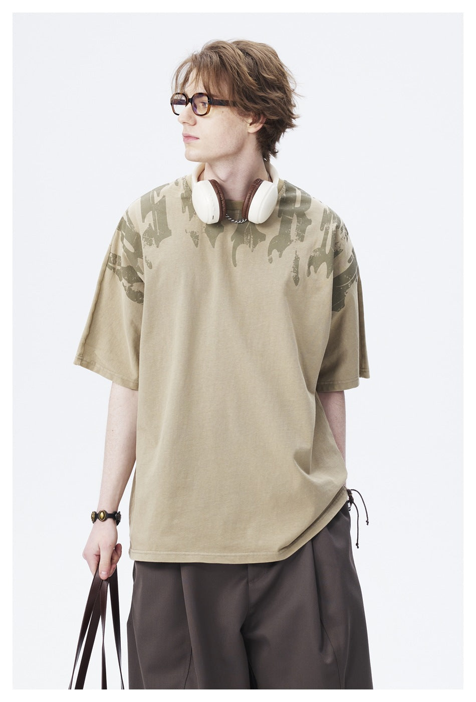 Model wearing an olive oversized T-shirt with dark abstract shoulder print and dark trousers.