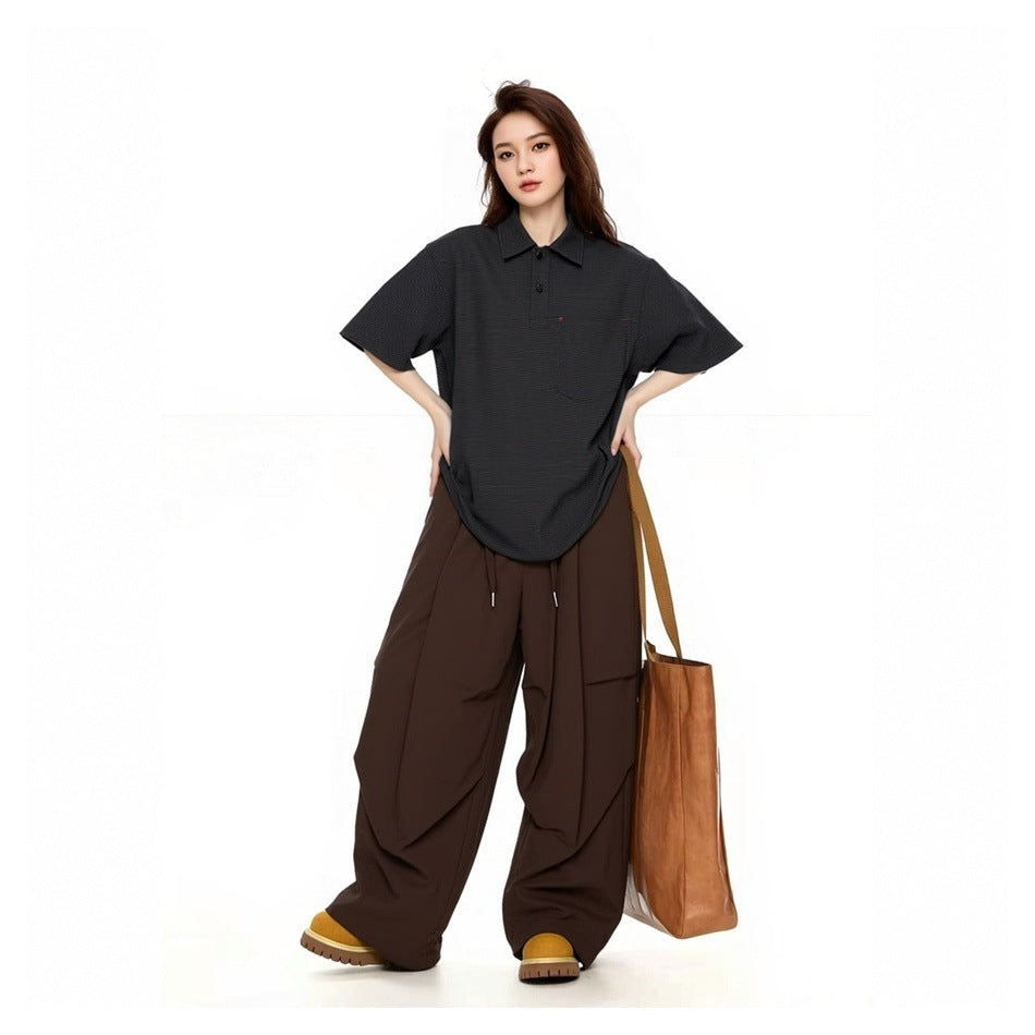 Model wearing an oversized black short-sleeve polo with brown wide-leg trousers and carrying a tan tote.