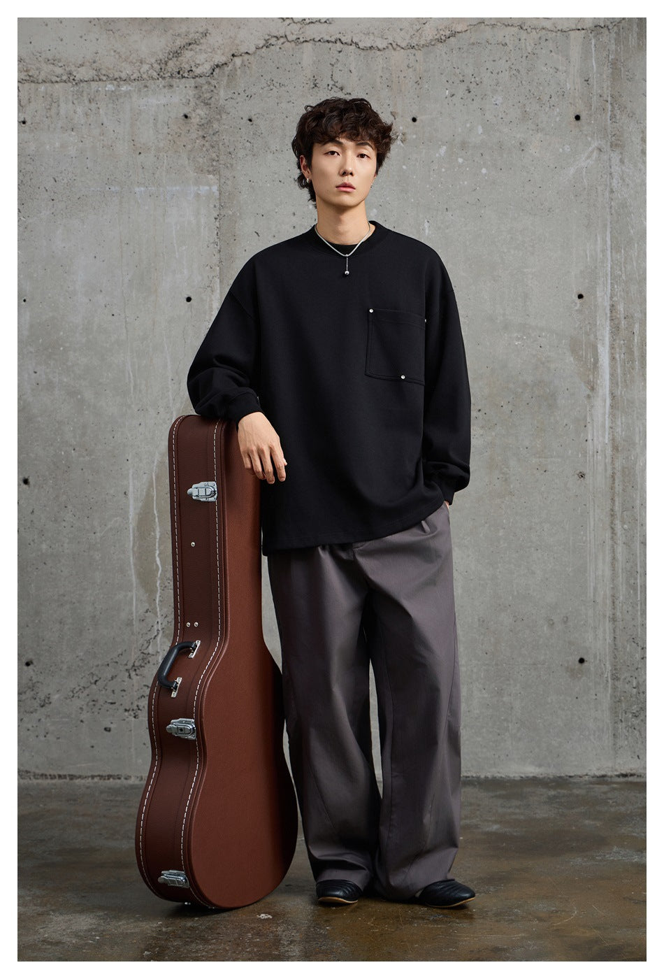 Front view of a model wearing a black oversized crewneck sweatshirt with a square chest pocket and wide gray trousers.
