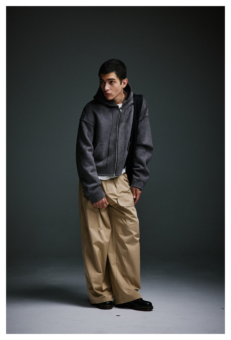 Full-length view of a model in a charcoal zip-up hoodie and wide taupe cargo trousers, posed against a dark backdrop.