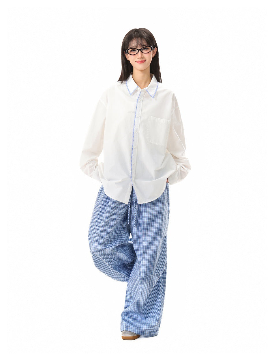 Full-length model wearing a white button-up shirt with blue collar trim and loose blue checked trousers.