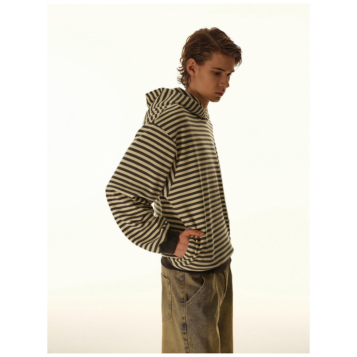 Side view of a model wearing a loose beige and dark striped hooded sweatshirt with dark ribbed cuffs and hem.