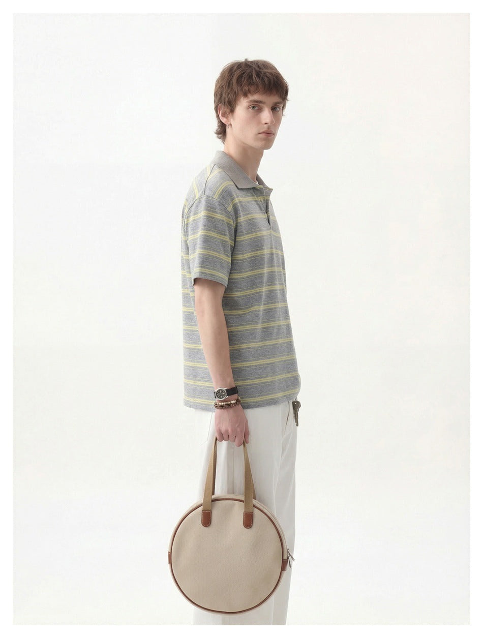 A man wears a gray polo with thin yellow stripes and white trousers, holding a round beige handbag.