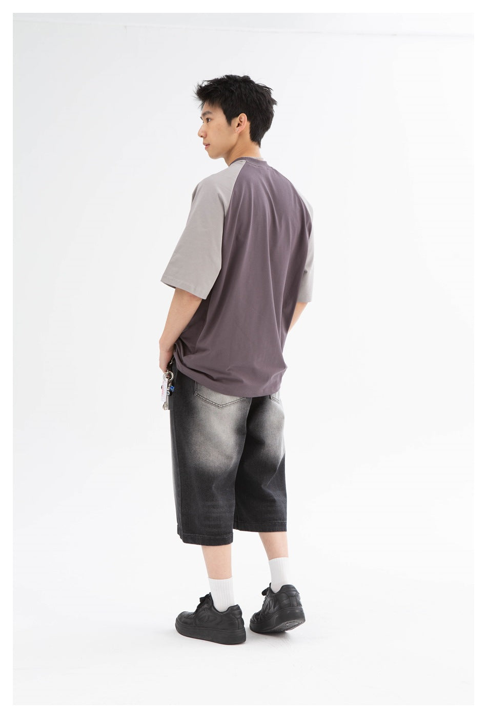Model seen from the back wearing black faded denim knee-length shorts with a gray and purple tee and black sneakers