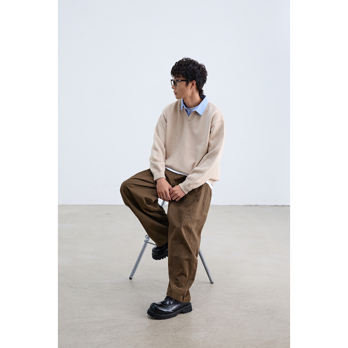 Seated model wearing a beige V-neck sweater over a blue striped shirt with brown trousers.