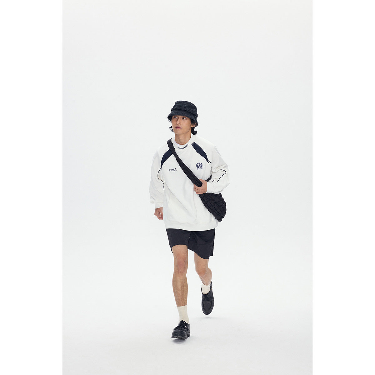 Full-length model wearing a white sweatshirt with navy accents and black shorts, walking with a black shoulder bag.