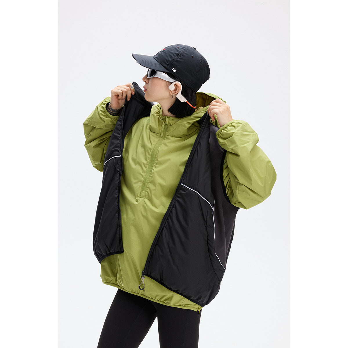Model opening a black sleeveless vest over an olive-green hooded pullover.