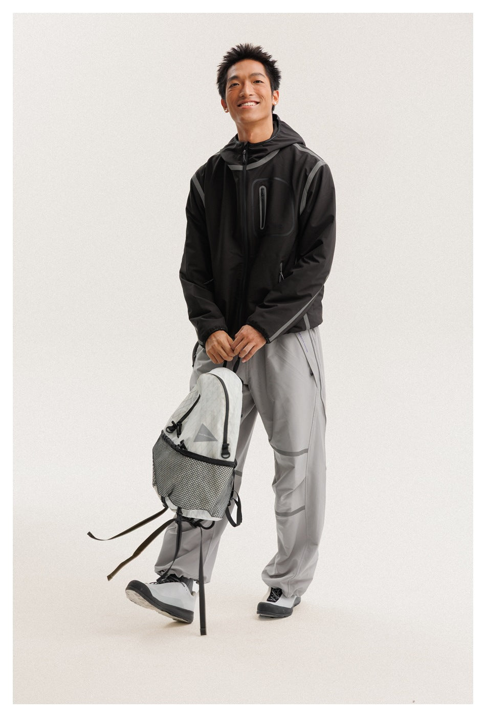 Model wearing a black hooded jacket with gray trim and zip pockets, paired with light gray trousers and carrying a backpack.