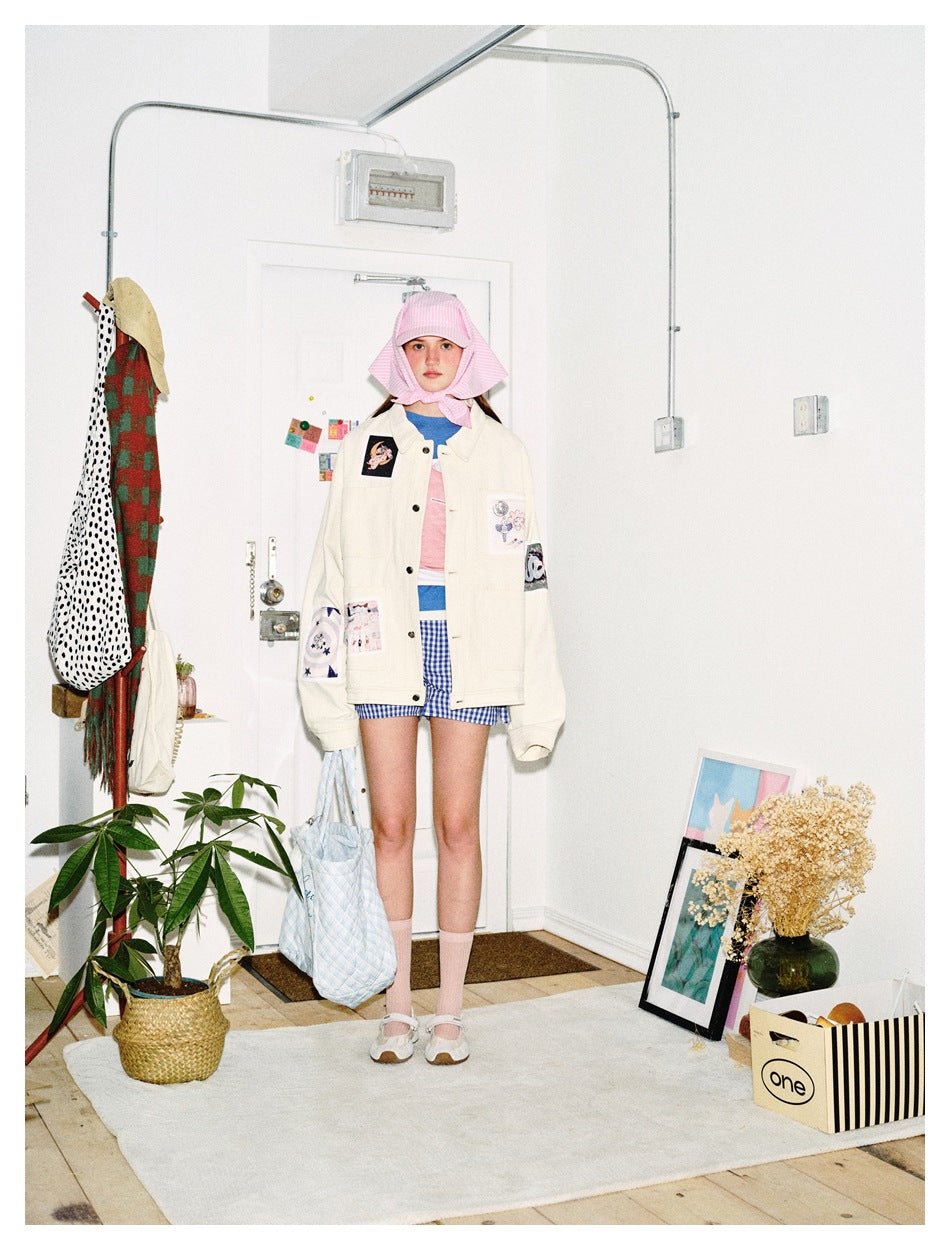 Model wearing a beige patch-detail jacket with pink headscarf and blue gingham shorts, front view, holding a quilted tote