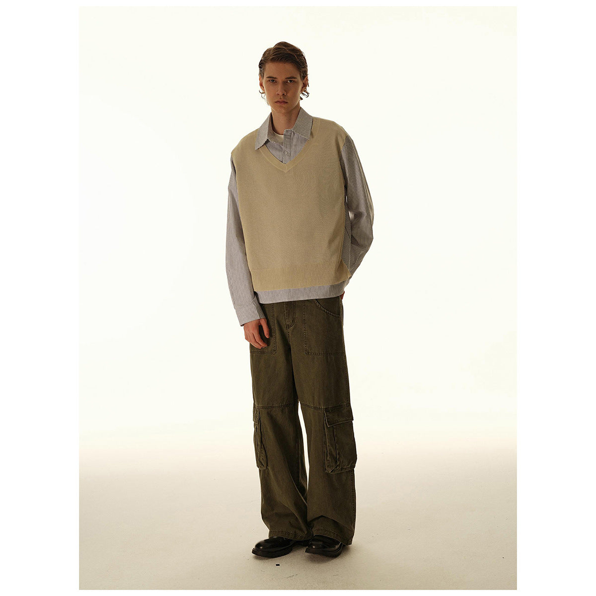 Three-quarter view of a model in a tan V-neck sweater vest, striped shirt, and dark olive cargo pants.