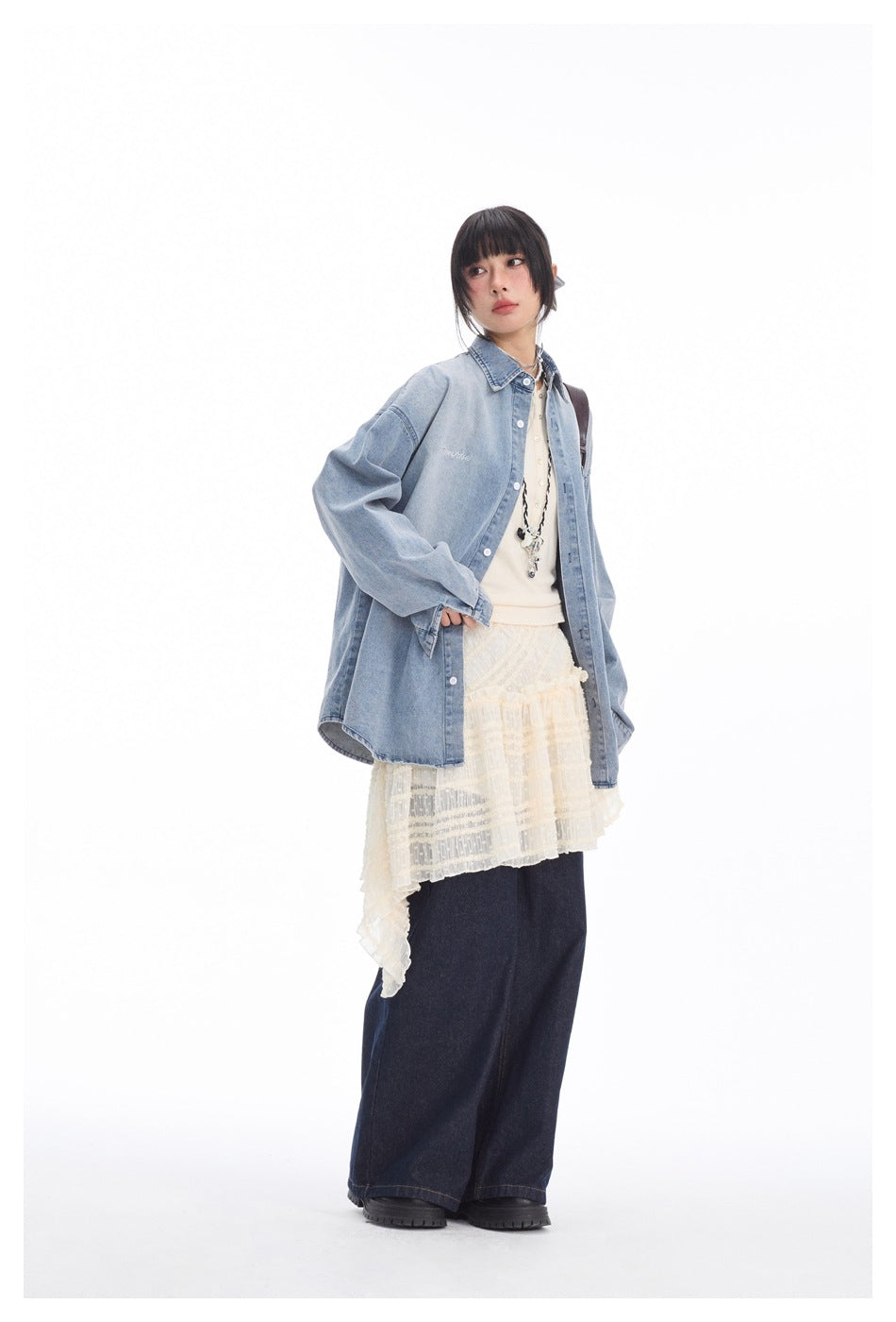 Person wearing an oversized light blue denim shirt open over a cream top, lace skirt and dark wide-leg jeans