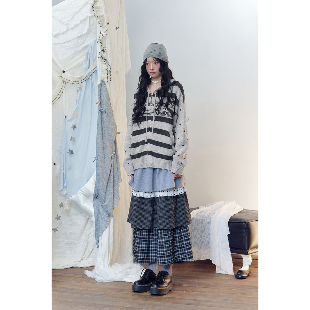 Full-length model wearing a gray striped hooded sweater, gray beanie, layered plaid skirt and chunky black shoes.