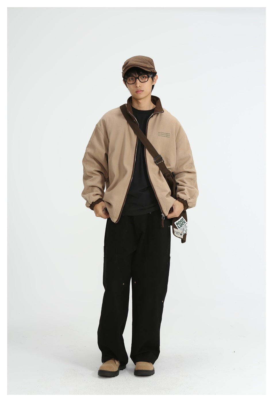 Model wearing a khaki zip jacket with brown collar over a black tee, black trousers, brown cap and a crossbody bag