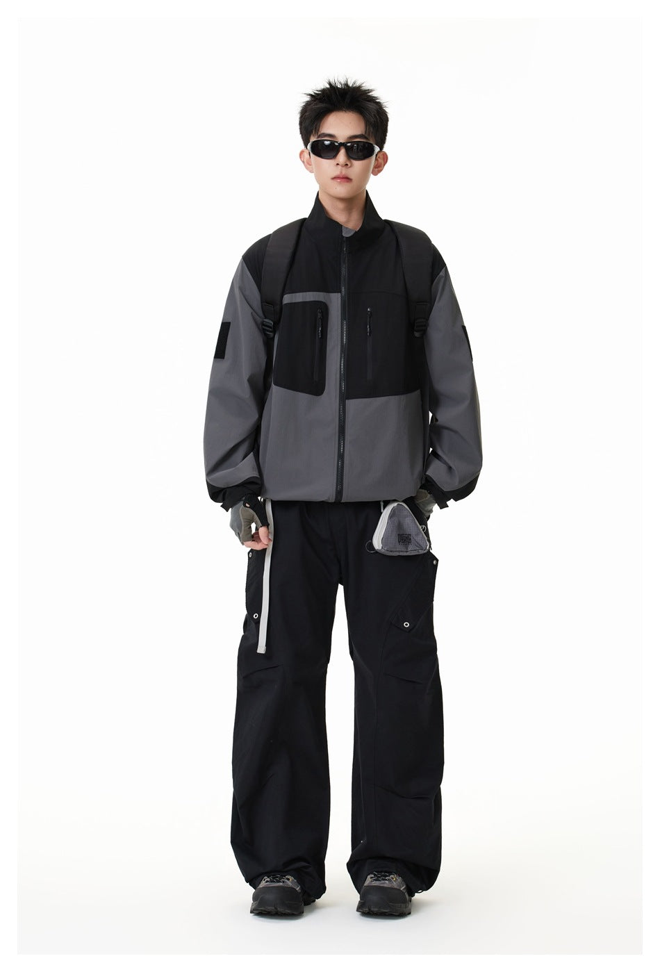 Full-length model wearing the black and charcoal-gray color-block jacket with black cargo pants and a backpack.