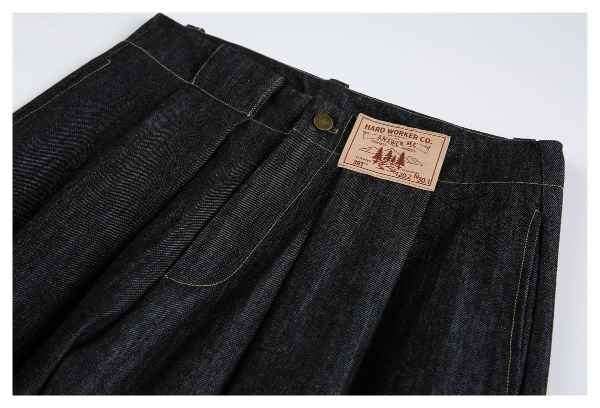 Upper-front close-up of dark indigo jeans with pleats, a brass button, pockets and a sewn tan patch.