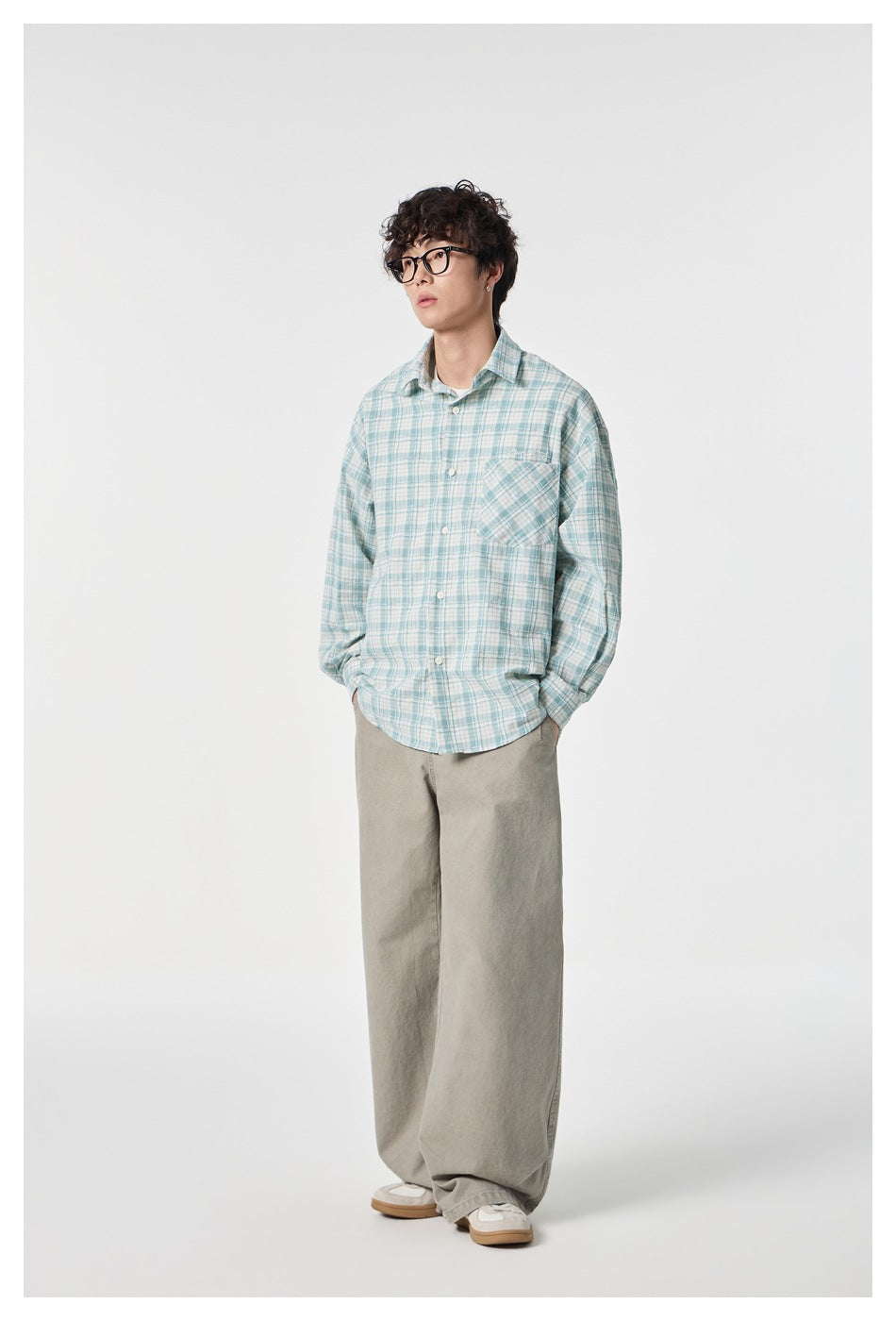 Man in a green plaid button-up shirt worn open-collared with wide beige pants, standing with hands in pockets