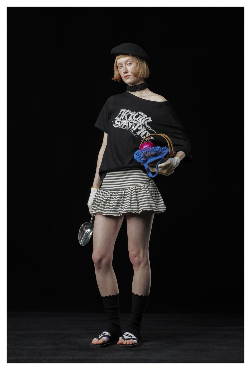 Model in black graphic T-shirt with slanted neckline, striped ruffle skirt, holding a basket and a scoop