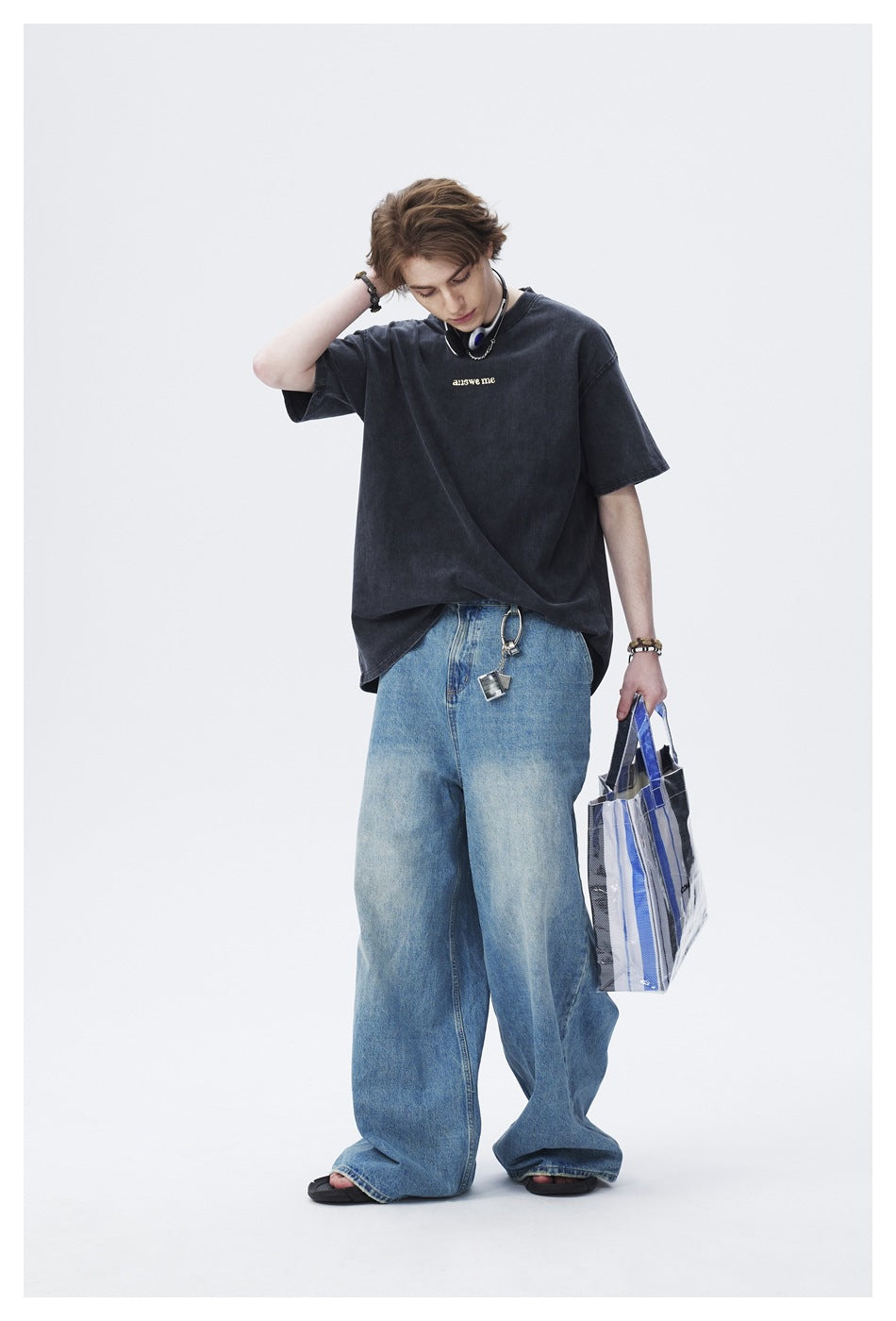 Model wearing a dark oversized T-shirt with small chest lettering and loose blue jeans, holding a striped tote.