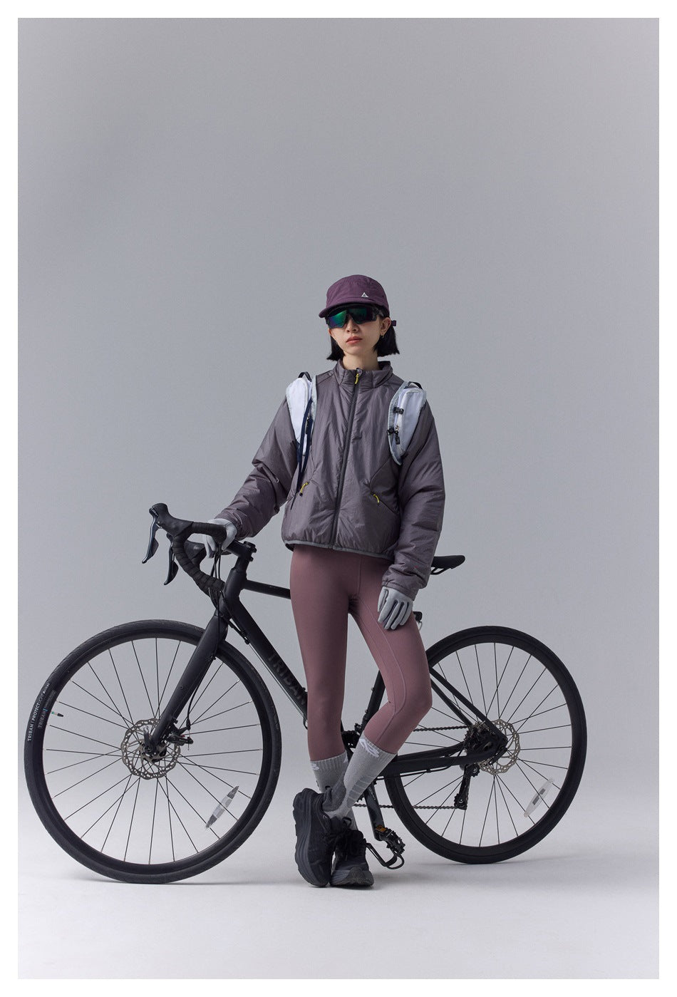 Model in a gray-plum zip jacket and mauve leggings standing beside a road bike, wearing a cap, sunglasses, gloves, and a