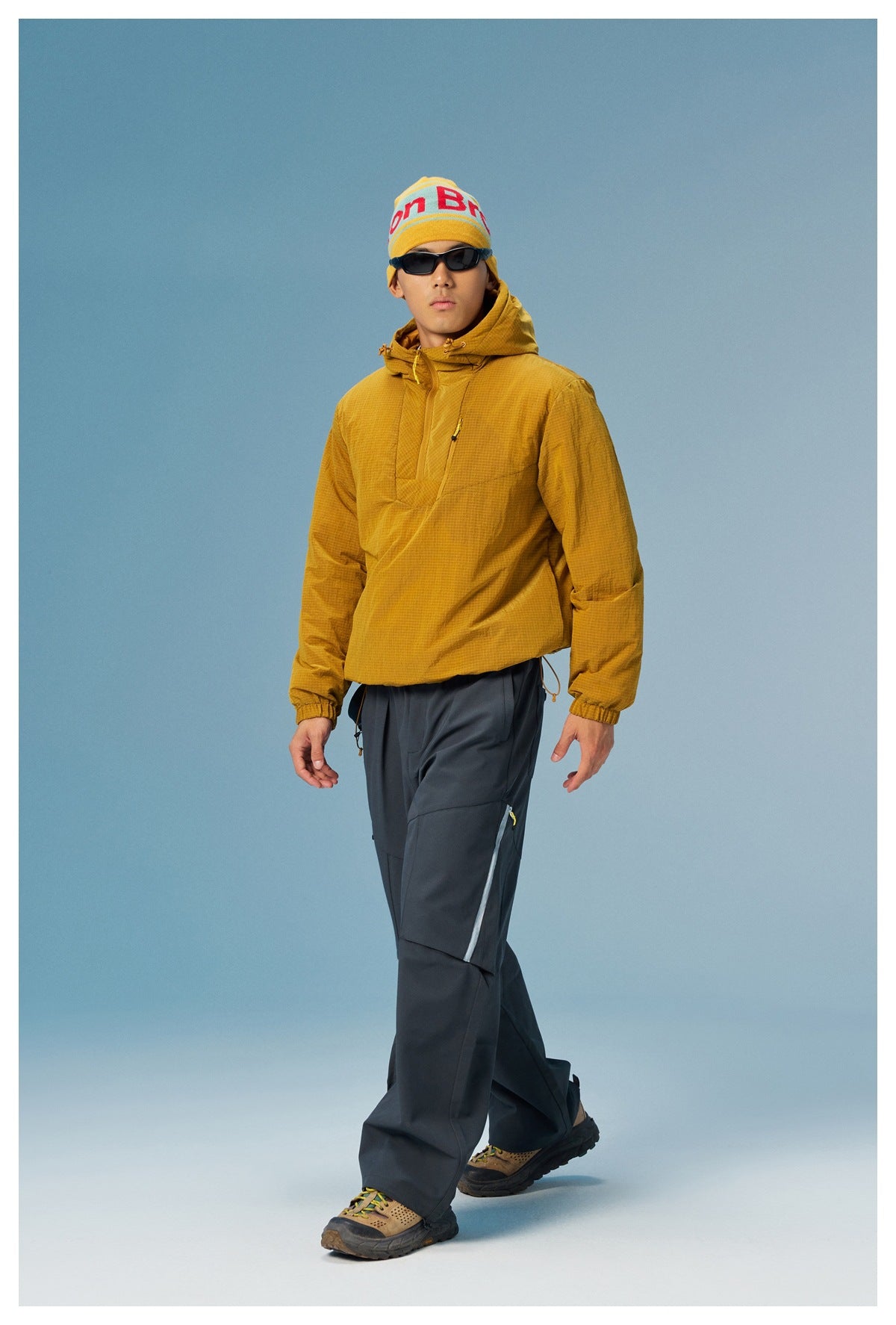 Man modeling a mustard grid-pattern hooded pullover with dark outdoor pants and a yellow knit beanie.