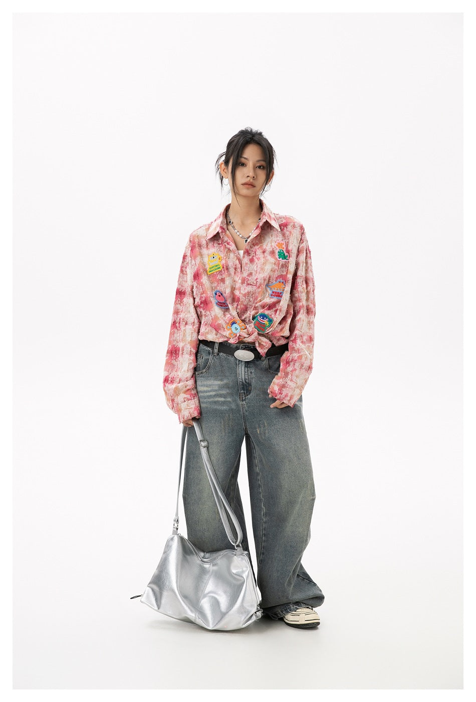 Full-length view of a model wearing a pink patterned button-up shirt tied at the waist with colorful patches and loose jeans.